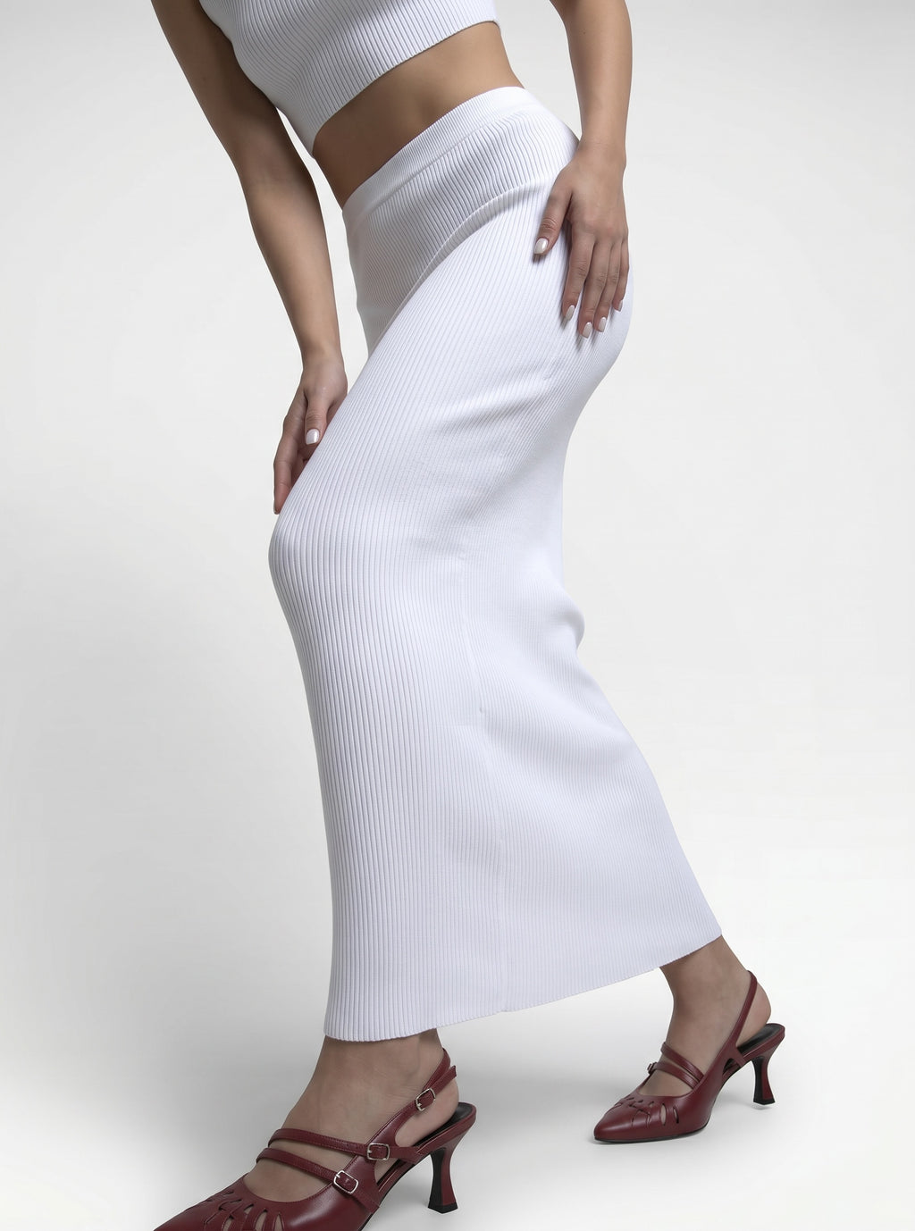 A model wearing a white ribbed knit midi skirt paired with a matching white crop top and dark red slingback kitten heels, shown from the waist down against a light gray studio background.