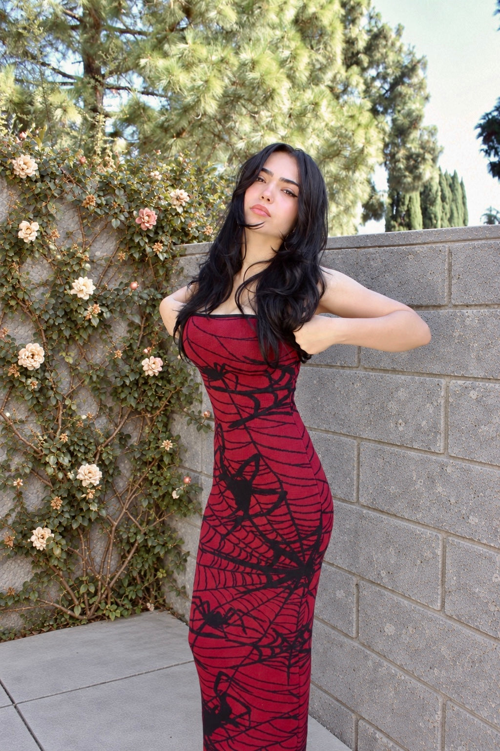 A woman posing outdoors in a red strapless maxi dress with a black spiderweb pattern, standing next to a gray brick wall and a flowering green vine.