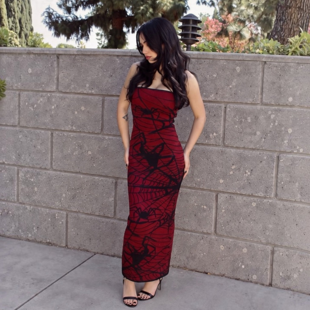 A woman wearing a strapless red maxi dress with a black spiderweb and spider print, posing outdoors against a gray concrete block wall.
