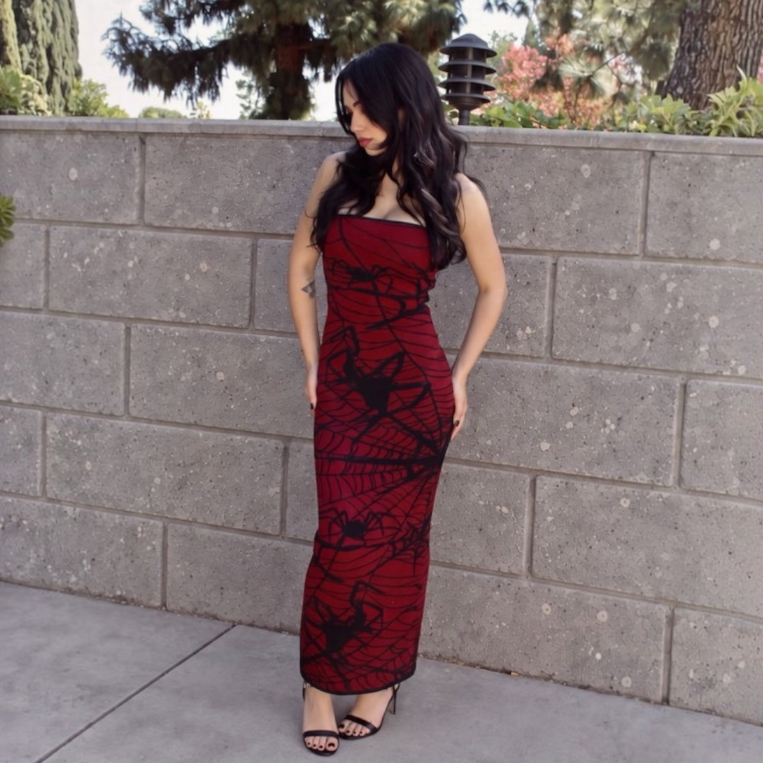 A woman wearing a strapless red maxi dress with a black spiderweb and spider print, posing outdoors against a gray concrete block wall.